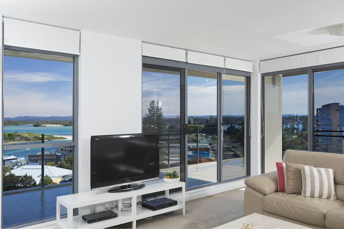 A modern living room with a large flat-screen TV on a white stand, a beige sofa with striped cushions, and large floor-to-ceiling windows offering a view of a body of water and distant buildings.