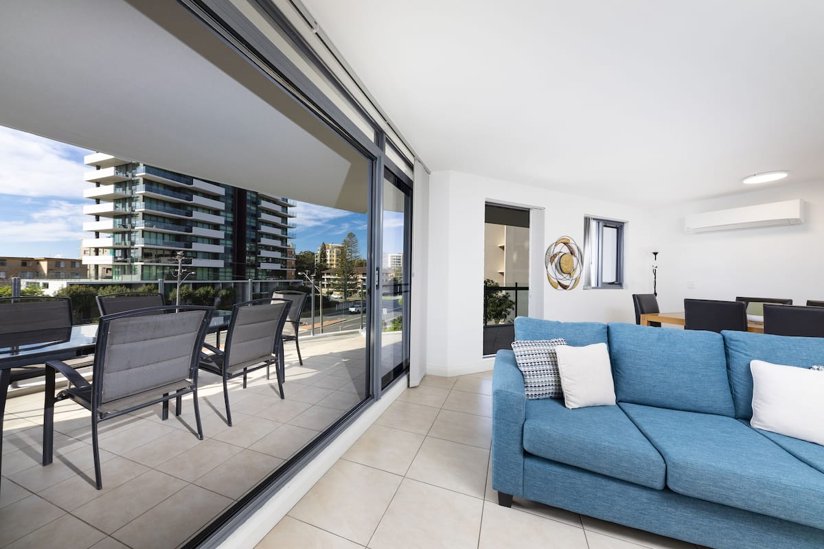 A modern living room with a blue sofa, a dining area with chairs, and a large sliding glass door leading to a balcony with a view of buildings.