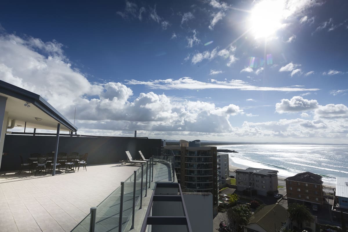 A rooftop terrace with a view of the ocean, a covered seating area, and a glass railing.