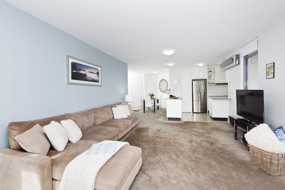 A living room with a beige sofa, a TV, and a kitchen in the background.