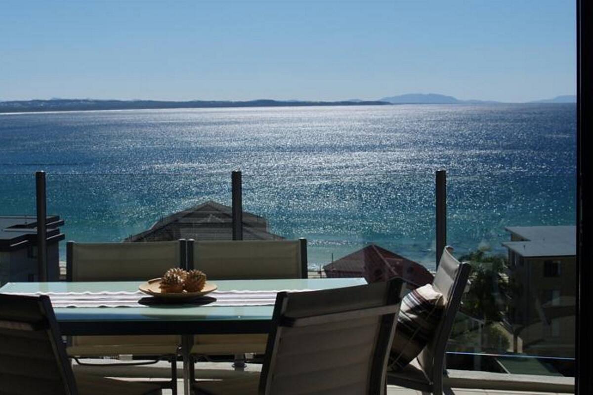 A table with a plate of food and chairs, overlooking a body of water with buildings in the background.