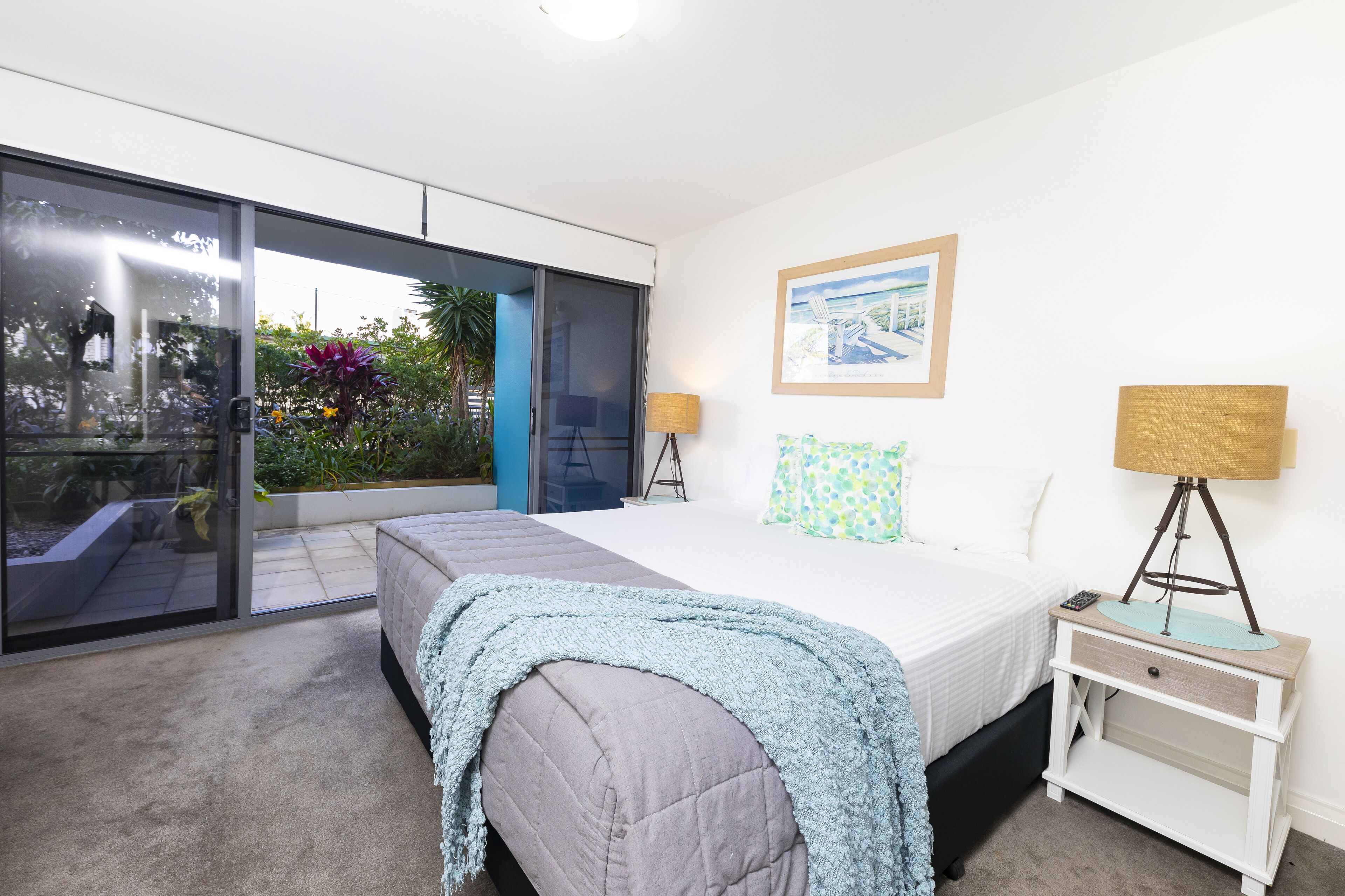 A bedroom with a bed, bedside table, lamp, and a view of the outdoors.