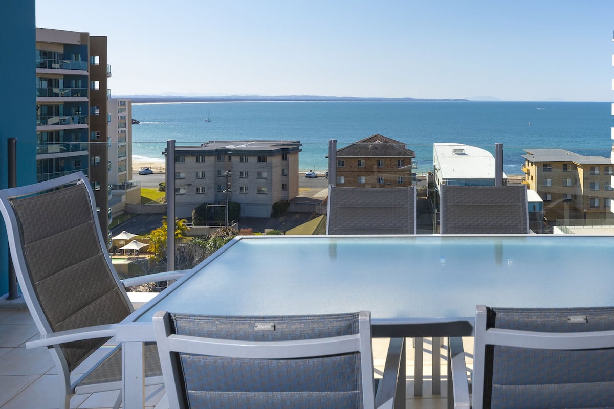 A balcony with a glass table and chairs overlooking a beach and ocean.