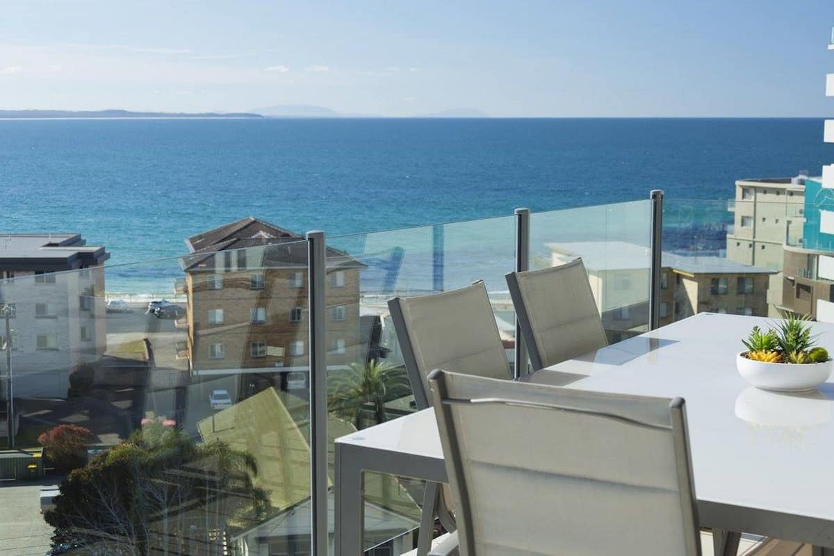 A balcony with a table and chairs, overlooking a coastal view with buildings and the sea.