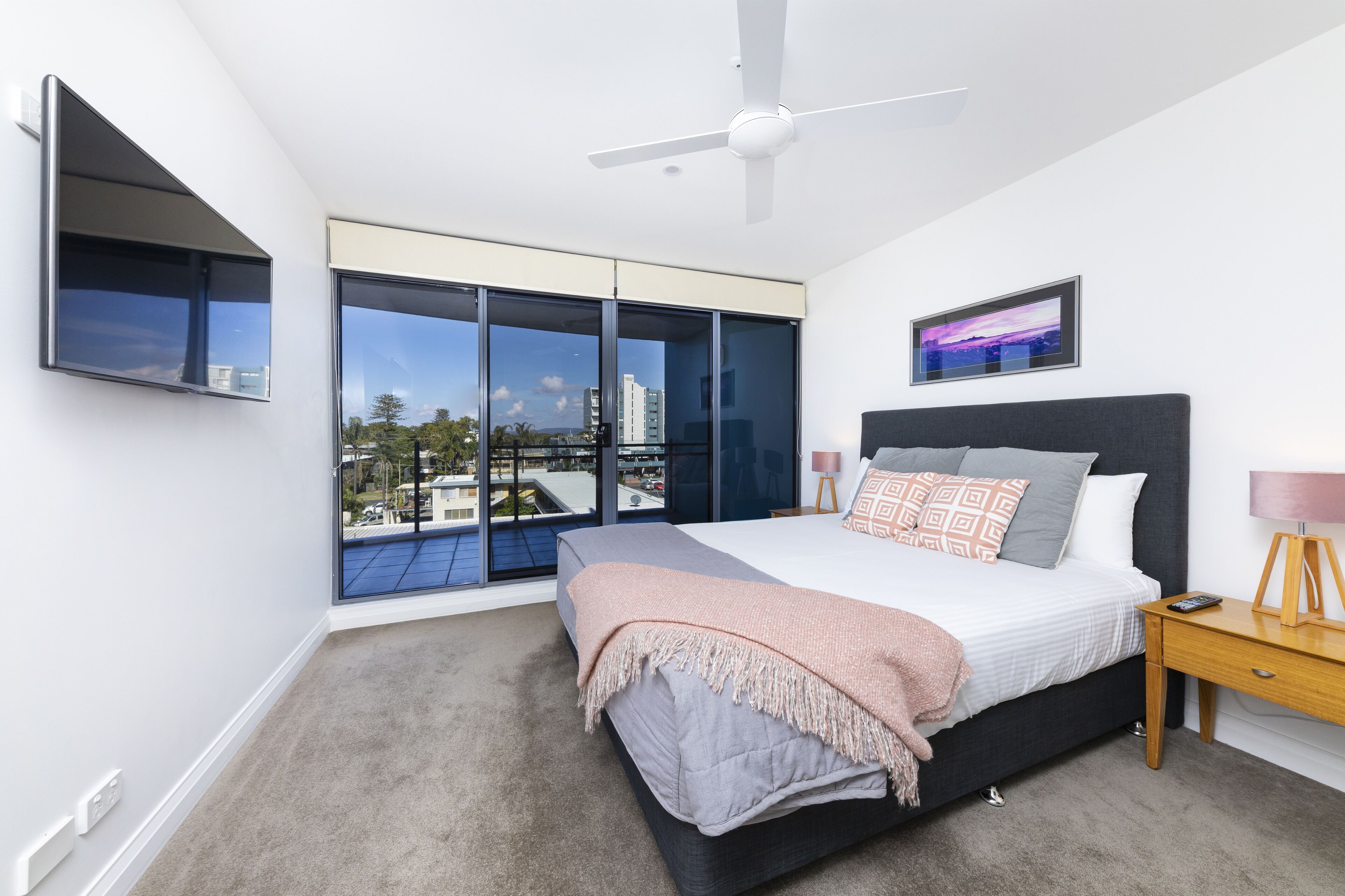 A modern bedroom with a large bed, a ceiling fan, a TV mounted on the wall, and a city view through the floor-to-ceiling windows.