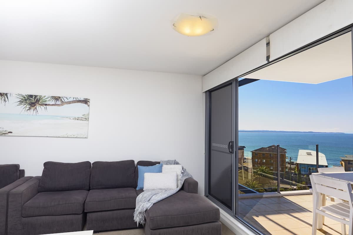 A modern living room with a grey sofa, a white chair, and a large window offering a view of the sea and buildings.