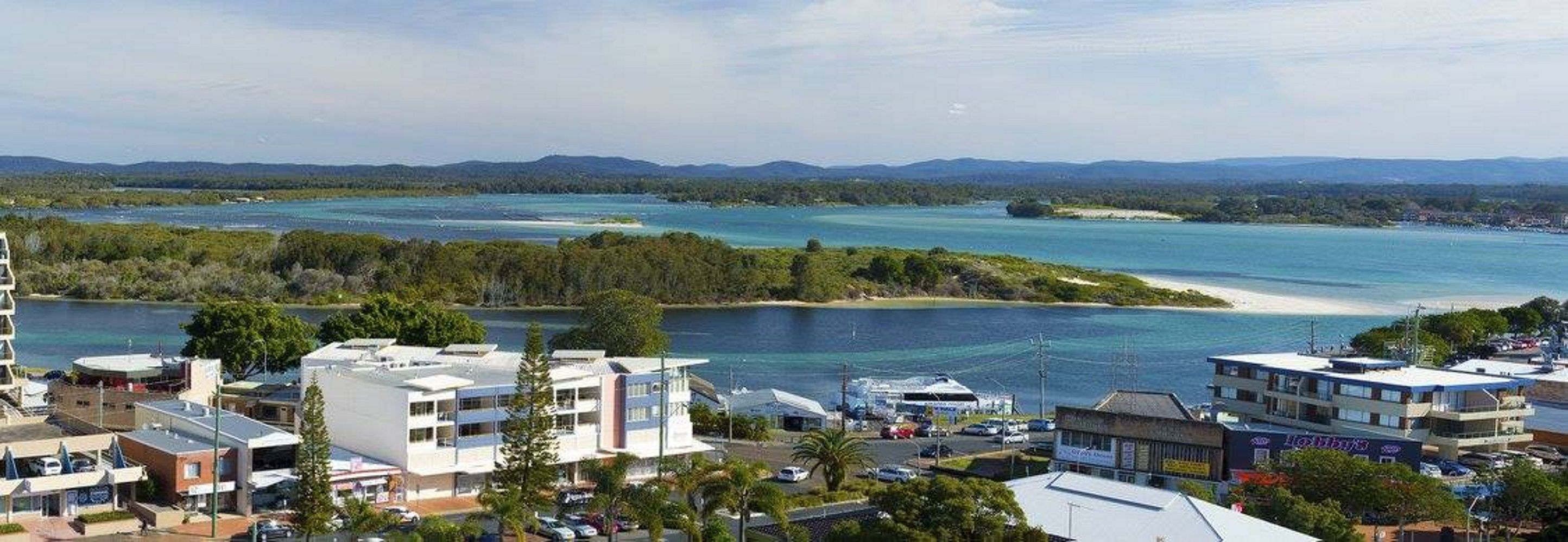 A coastal town with buildings near a large body of water, surrounded by greenery and hills in the background.