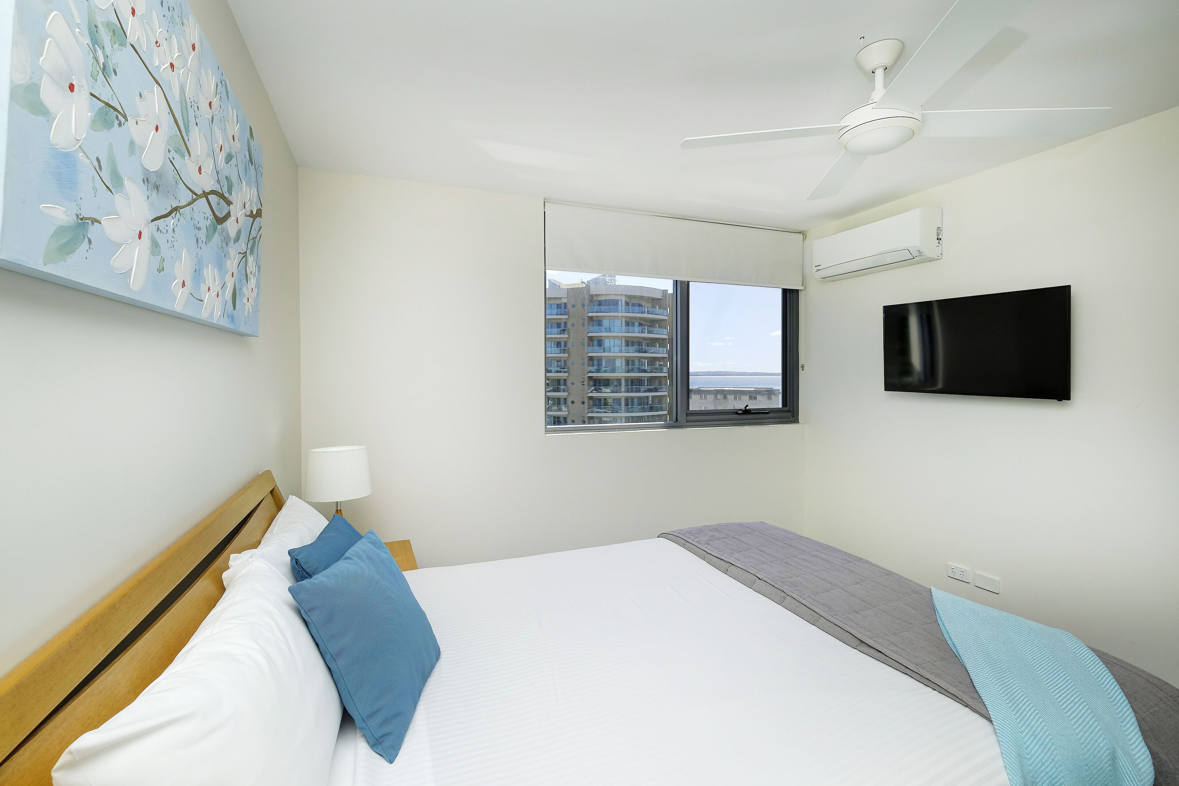 A hotel room with a bed, a ceiling fan, a TV, and a window with a view of buildings.