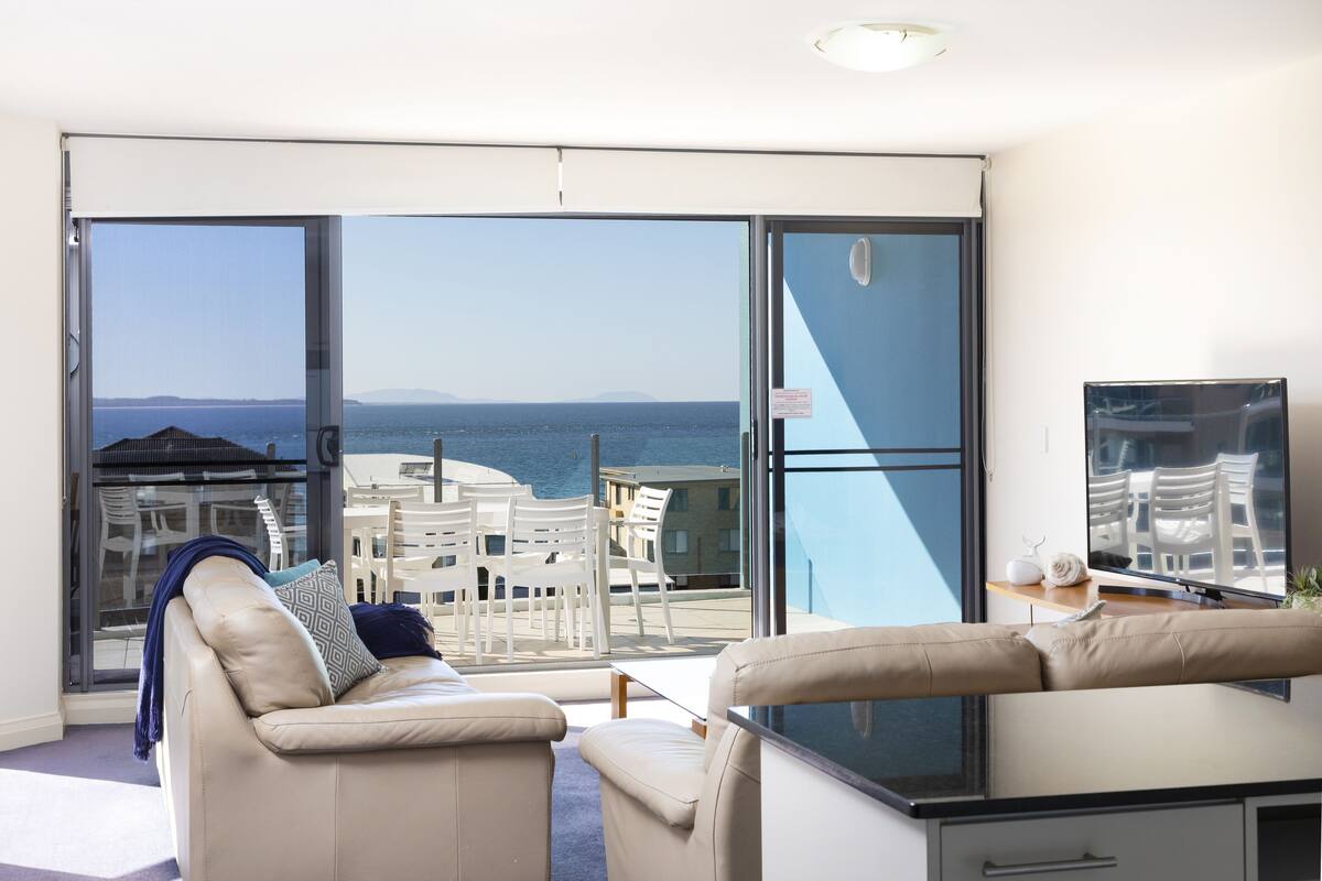 A modern living room with a sofa, a TV, and a balcony with a view of the sea.