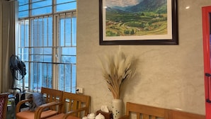 A room with a large window, a framed landscape painting, a fan, a vase with dried plants, and wooden furniture. - A25 Hotel - Doi Can 2 (Hanoi)