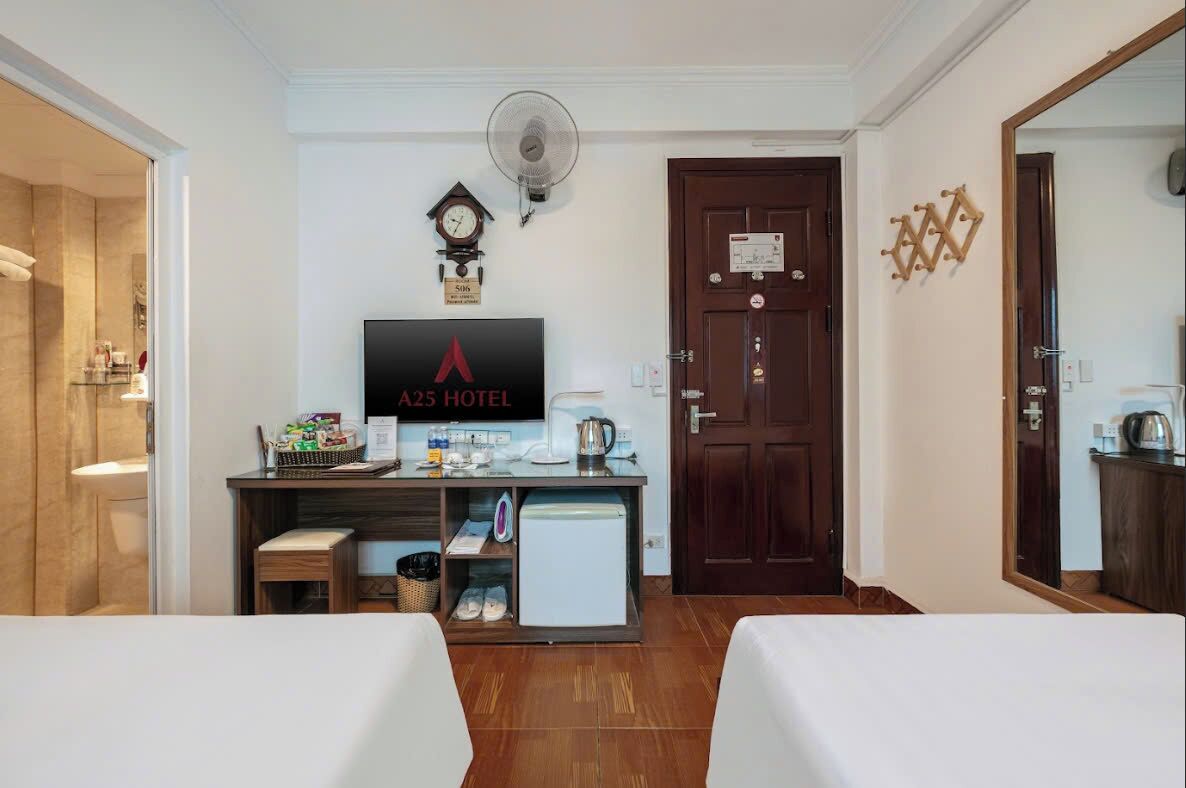 Habitación de hotel con dos camas, un escritorio con televisor mostrando el nombre del hotel, un reloj, un ventilador y una puerta con cerradura de tarjeta.