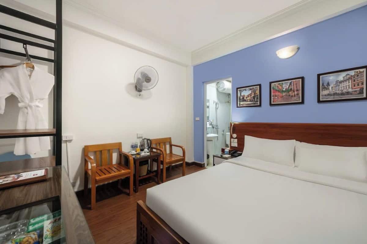 Habitación de hotel con cama, escritorio con sillas, ventilador y perchero con toallas.