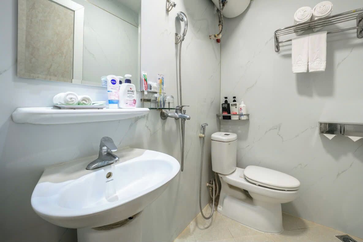 A bathroom with a sink, toilet, and shower. There are toiletries on the sink and a towel rack above the toilet.