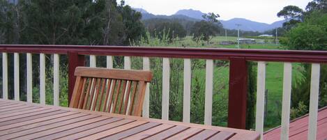 Una terraza de madera con barandal rojo, una mesa de madera y vista a un paisaje verde con árboles y colinas al fondo.