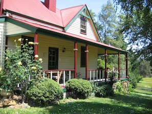 Una casa de dos pisos con un porche, techo rojo y jardín con arbustos y flores.