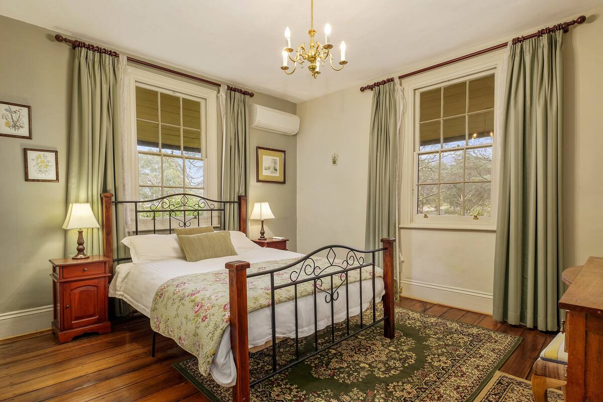 Un dormitorio con cama con dosel, dos mesitas de noche con lámparas, un candelabro y un ventanal con cortinas.