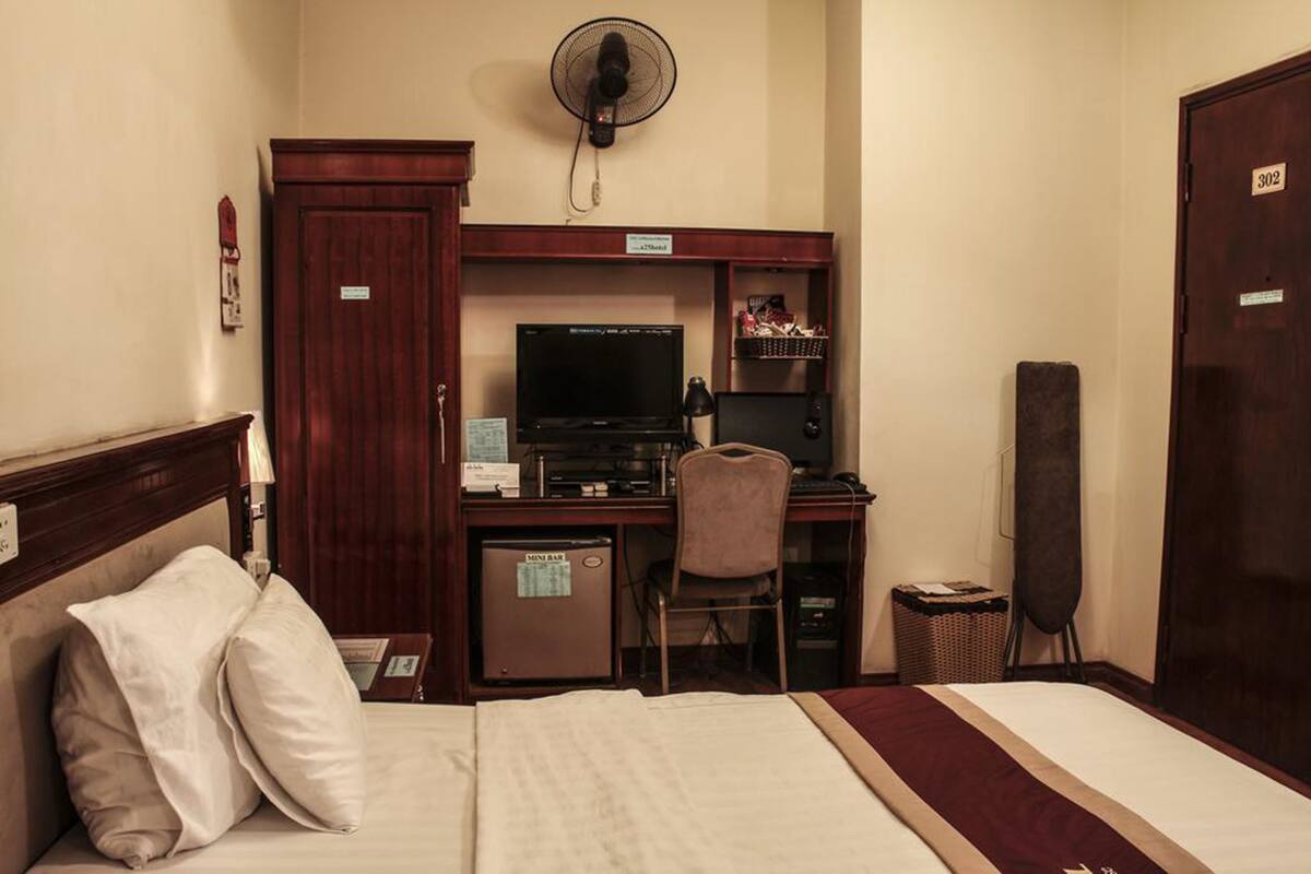 Een hotelkamer met een bed, een bureau met een computer, een stoel, een ventilator en een minibar.