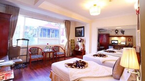 A hotel room with two beds, a desk, a chair, a window with curtains, and a small kitchenette.