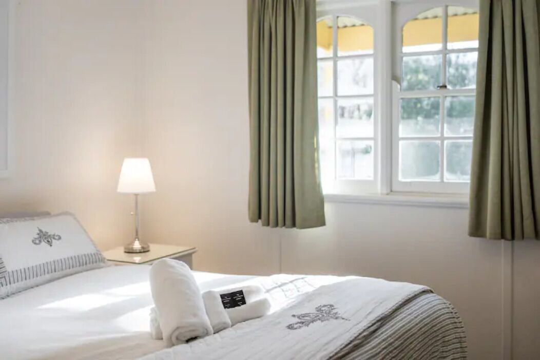 A neatly made bed with a white comforter and a decorative pillow, a bedside lamp, and a window with curtains.