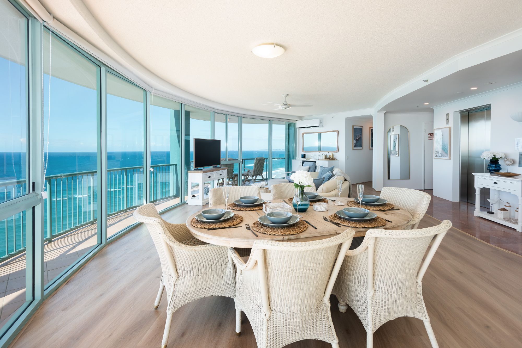Une pièce spacieuse avec une grande table à manger dressée pour six personnes, entourée de chaises en osier, d’un téléviseur et offrant une vue sur l’océan.