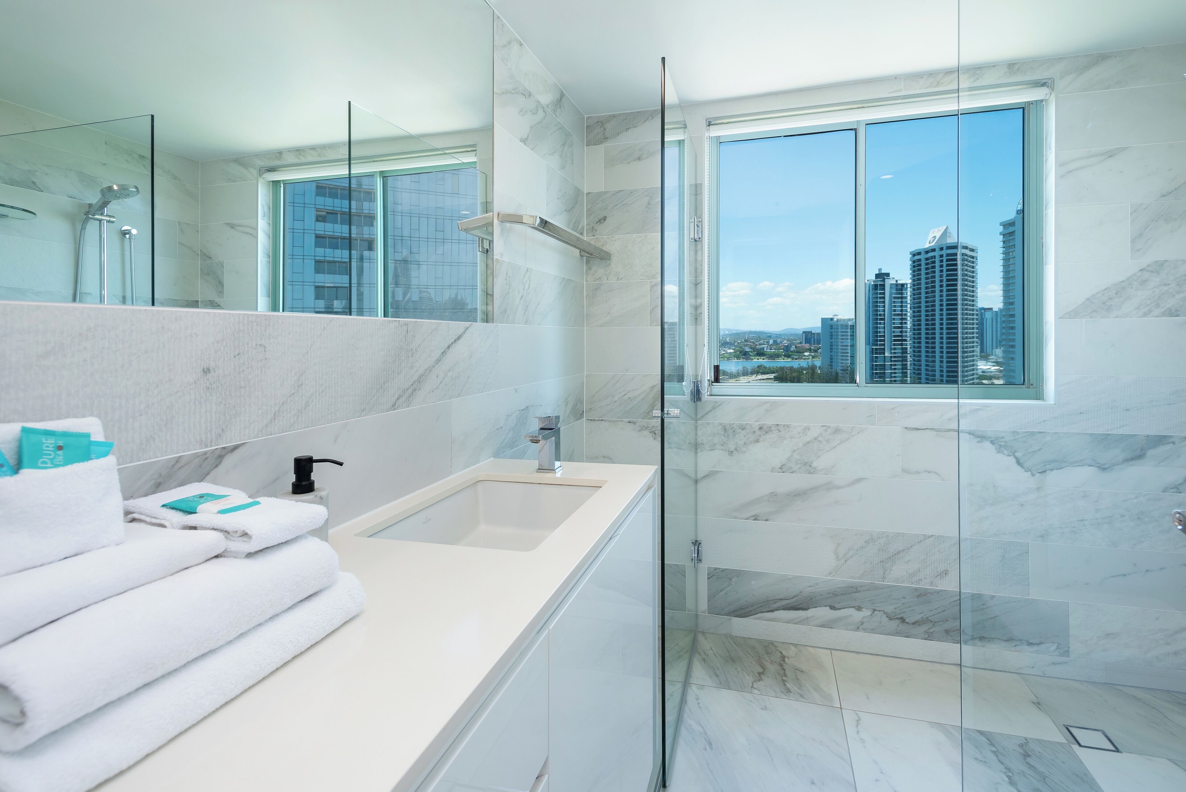 Un baño moderno con un espejo grande, ducha con mampara de vidrio, lavamanos blanco y vistas a un paisaje urbano a través de un ventanal amplio.