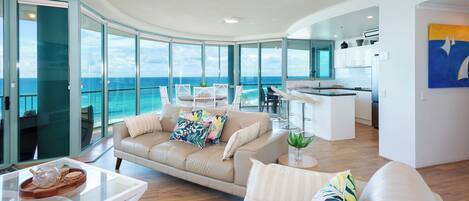 A spacious living area with a large window overlooking the ocean, a modern kitchen, and a seating area with a white sofa and coffee table.