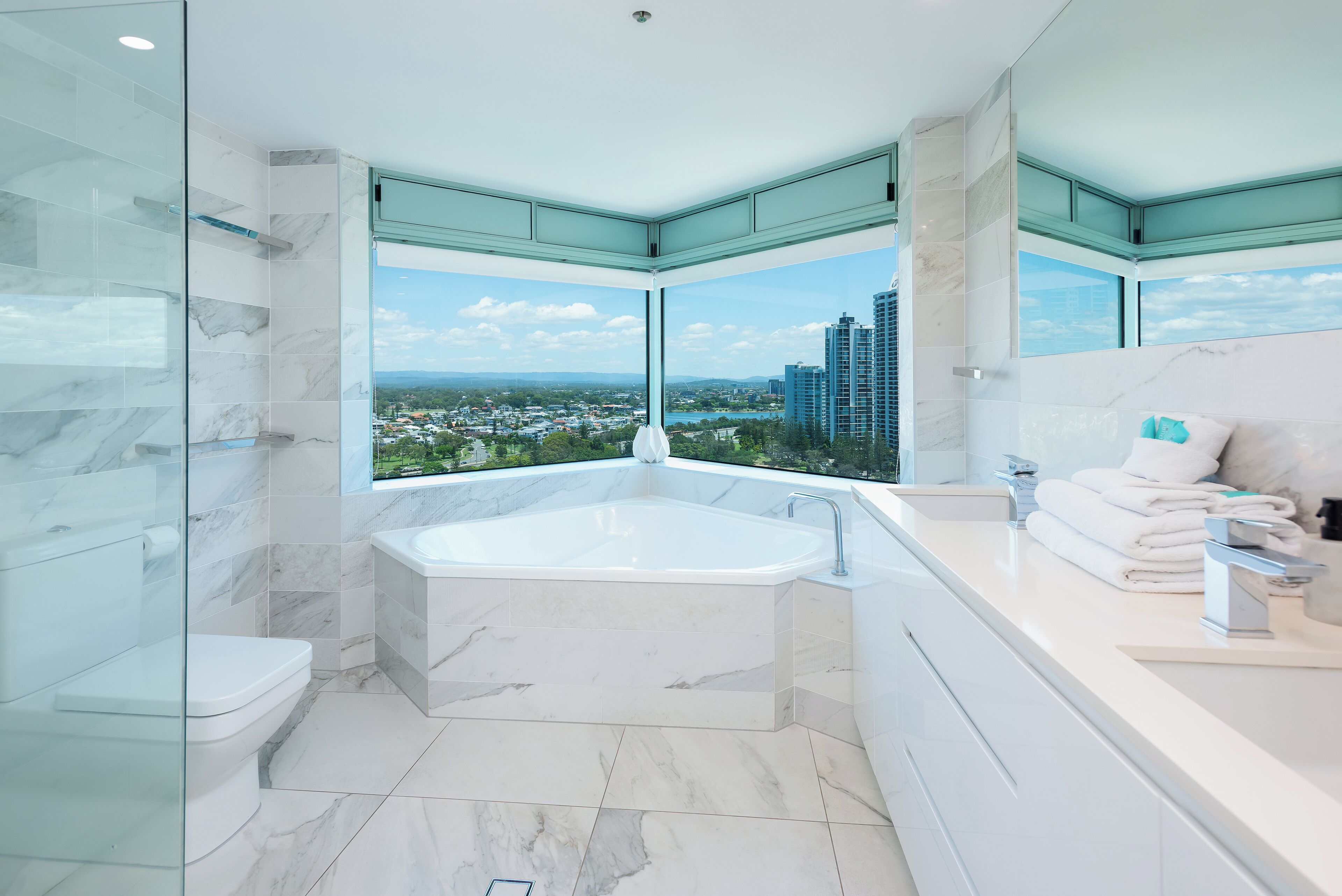 A modern bathroom with a large bathtub, a toilet, and a sink. There are large windows offering a view of a cityscape.