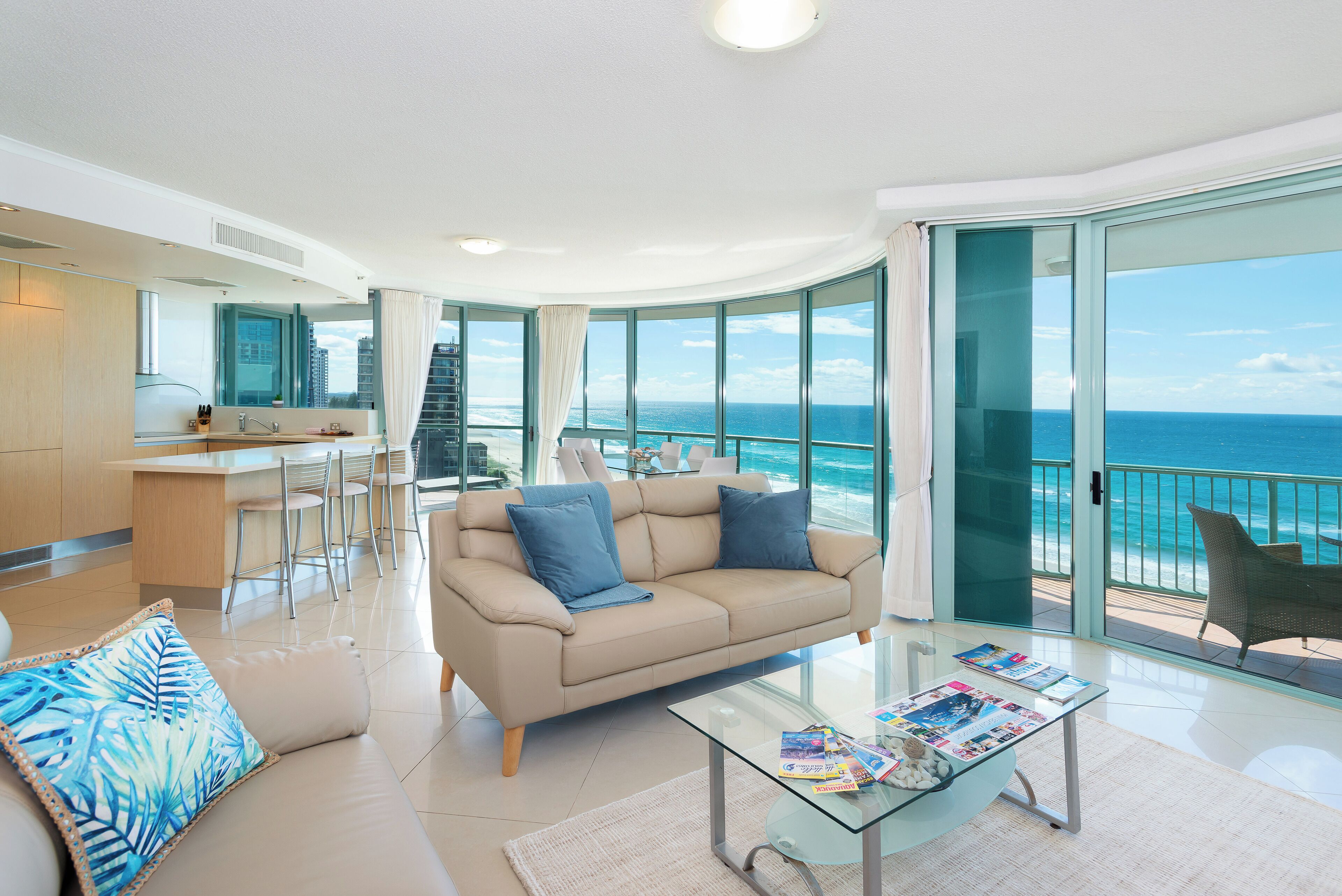 A modern living room with a beige sofa, a glass coffee table, and a view of the ocean.