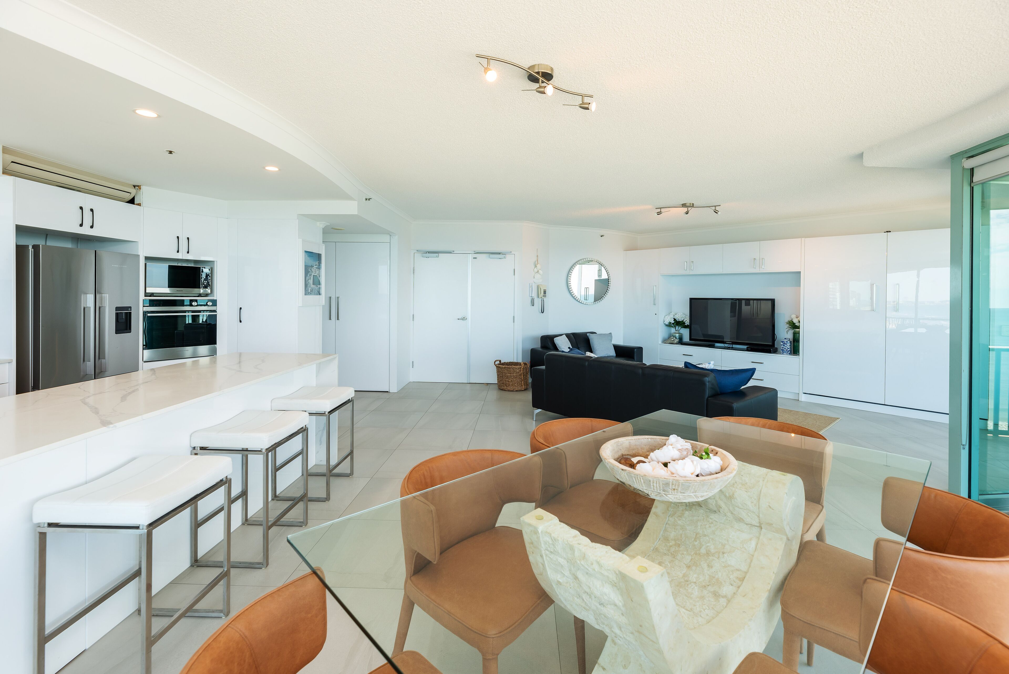 A modern kitchen with built-in appliances, a dining area with glass-top table and orange chairs, and a living area with a black sofa and TV.