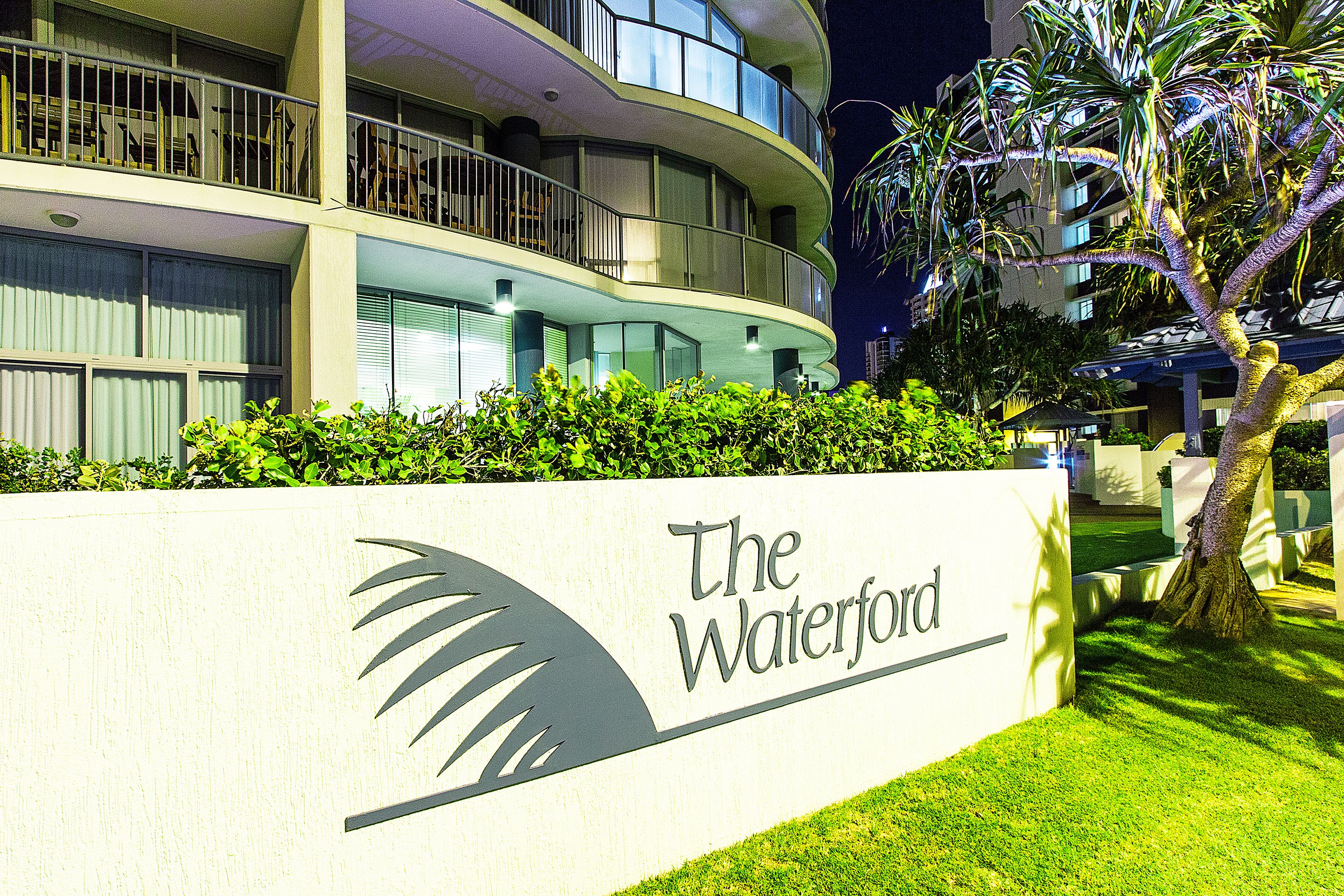 A modern hotel with a curved balcony, a sign reading "The Waterford," and well-maintained greenery.