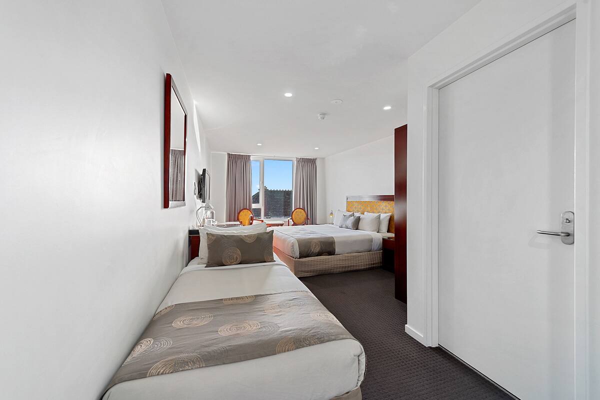 A hotel room with two beds, a large mirror, a desk with a chair, and a view of the cityscape through the window.