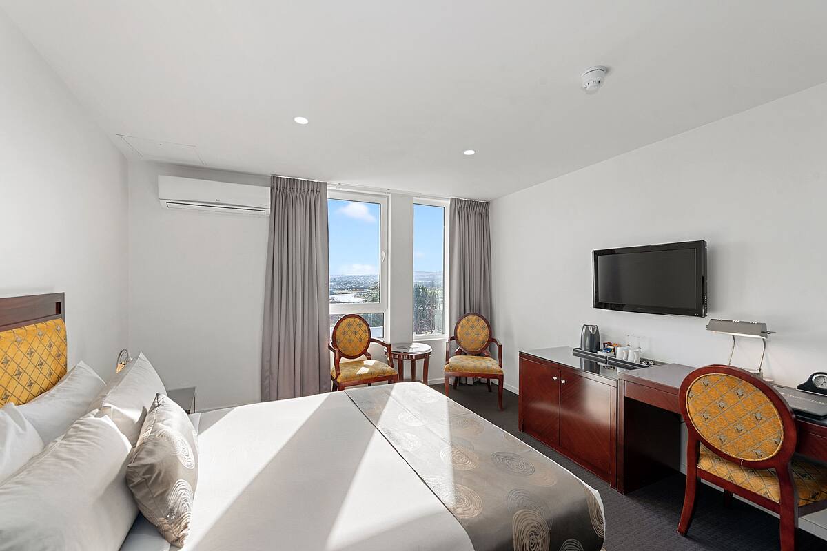 A hotel room with a bed, two chairs, a desk, a television, and a window with curtains.