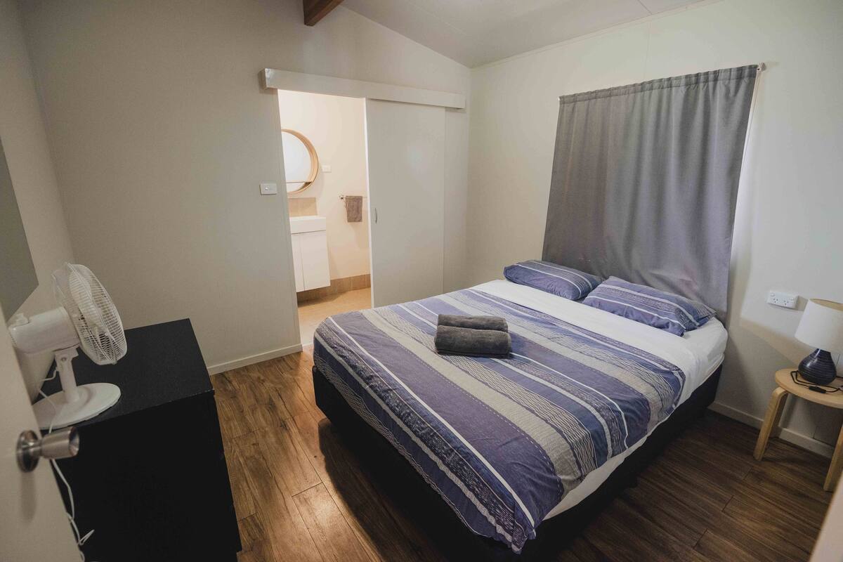 Un dormitori amb llit, ventilador, taula petita i porta que dona a un bany.