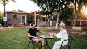 To personer spiser ved et udendørs bord med udsigt til en bygning, der er mærket "Camp Kitchen".