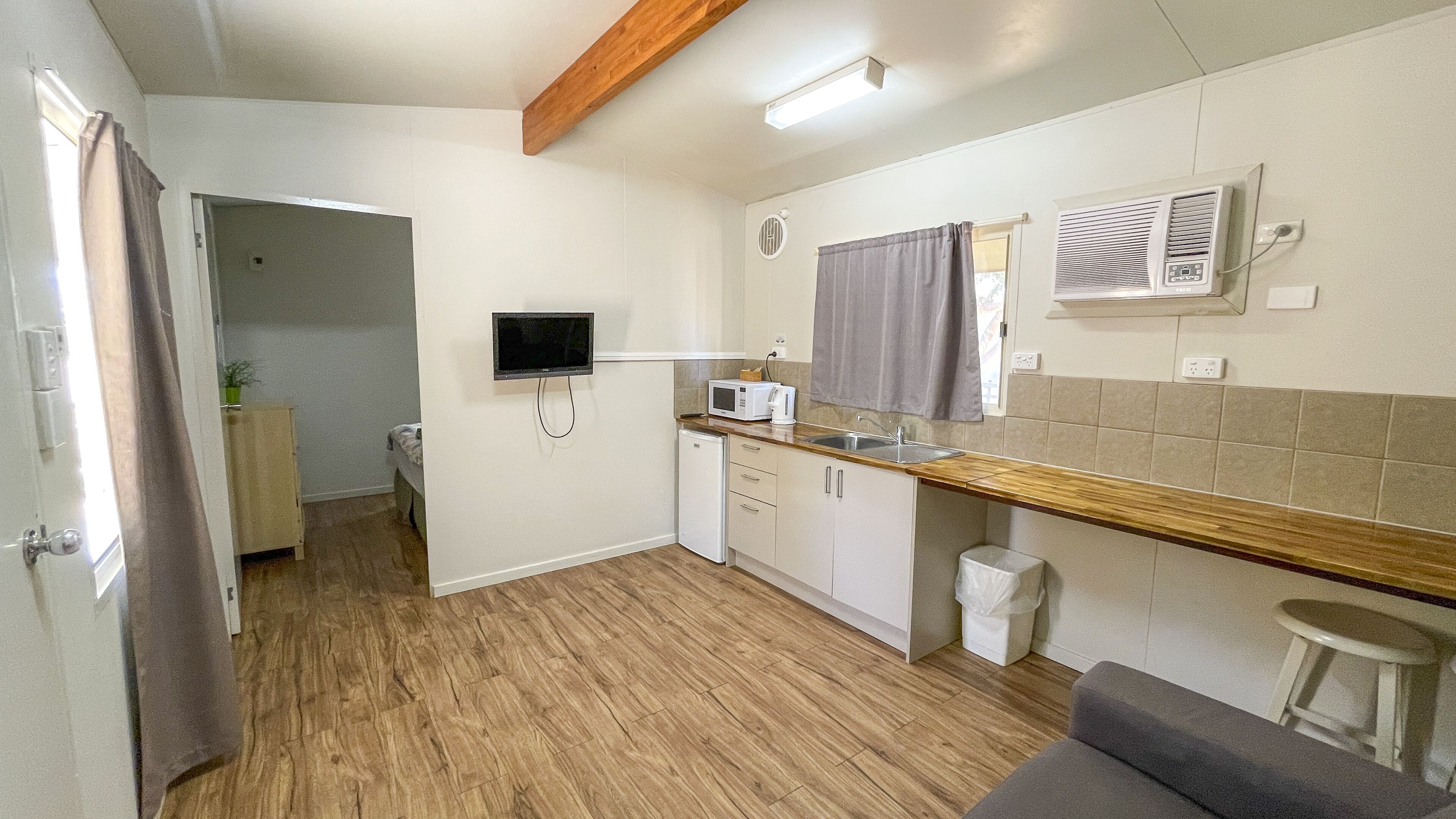 Una cocina compacta con microondas, pileta y heladera pequeña. Se observa una viga de techo de madera y un televisor de pantalla plana montado en la pared.