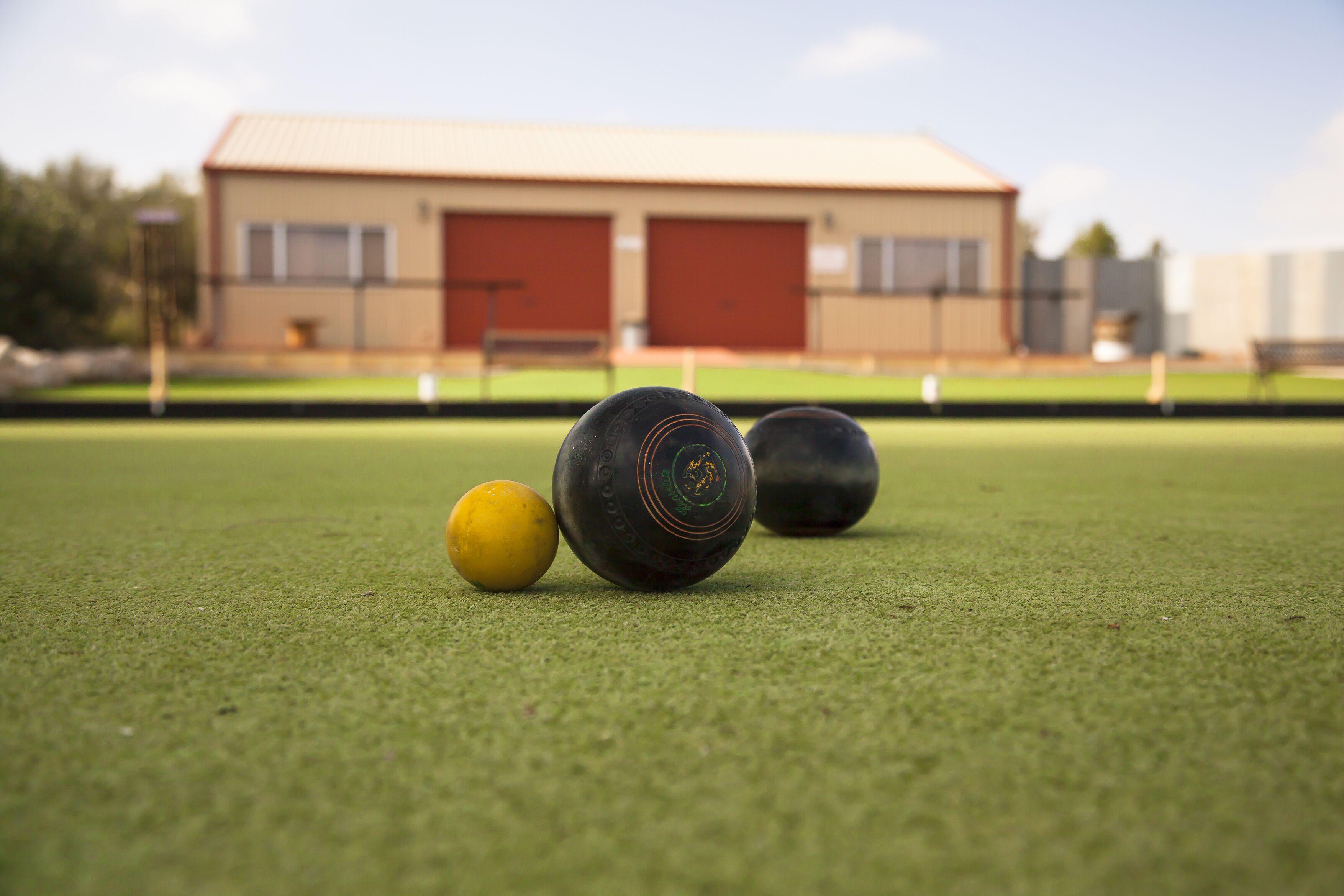 Primer plano de un césped con una pelota de croquet y una pelota amarilla, y un edificio con puertas rojas al fondo.