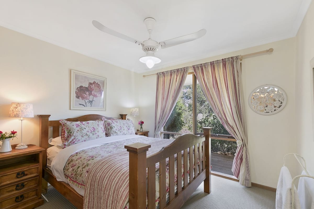 Un dormitorio con cama de madera, ventilador de techo, ventana con cortinas, mesita de noche con lámpara y un cuadro en la pared.