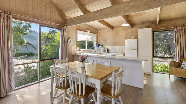 A wooden cabin interior with a dining area, kitchen, and large windows. - Freshwater Creek Cottages and Farm Stay (Freshwater Creek)