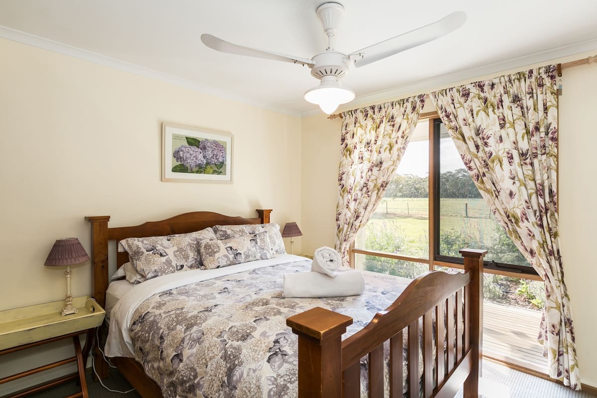 Un dormitorio con cama de madera, cortinas florales, ventilador de techo y ventana con vistas a zonas verdes.