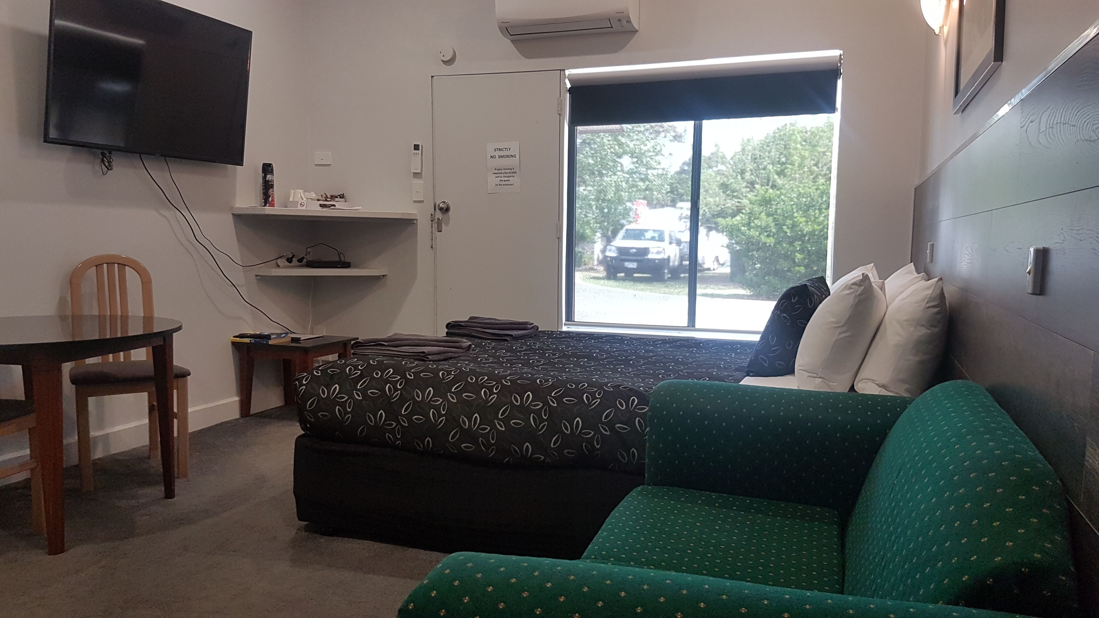 Ein Zimmer mit einem Bett, einem grünen Sofa, einem kleinen Tisch, einem an der Wand montierten Fernseher und einem Fenster mit Ausblick.