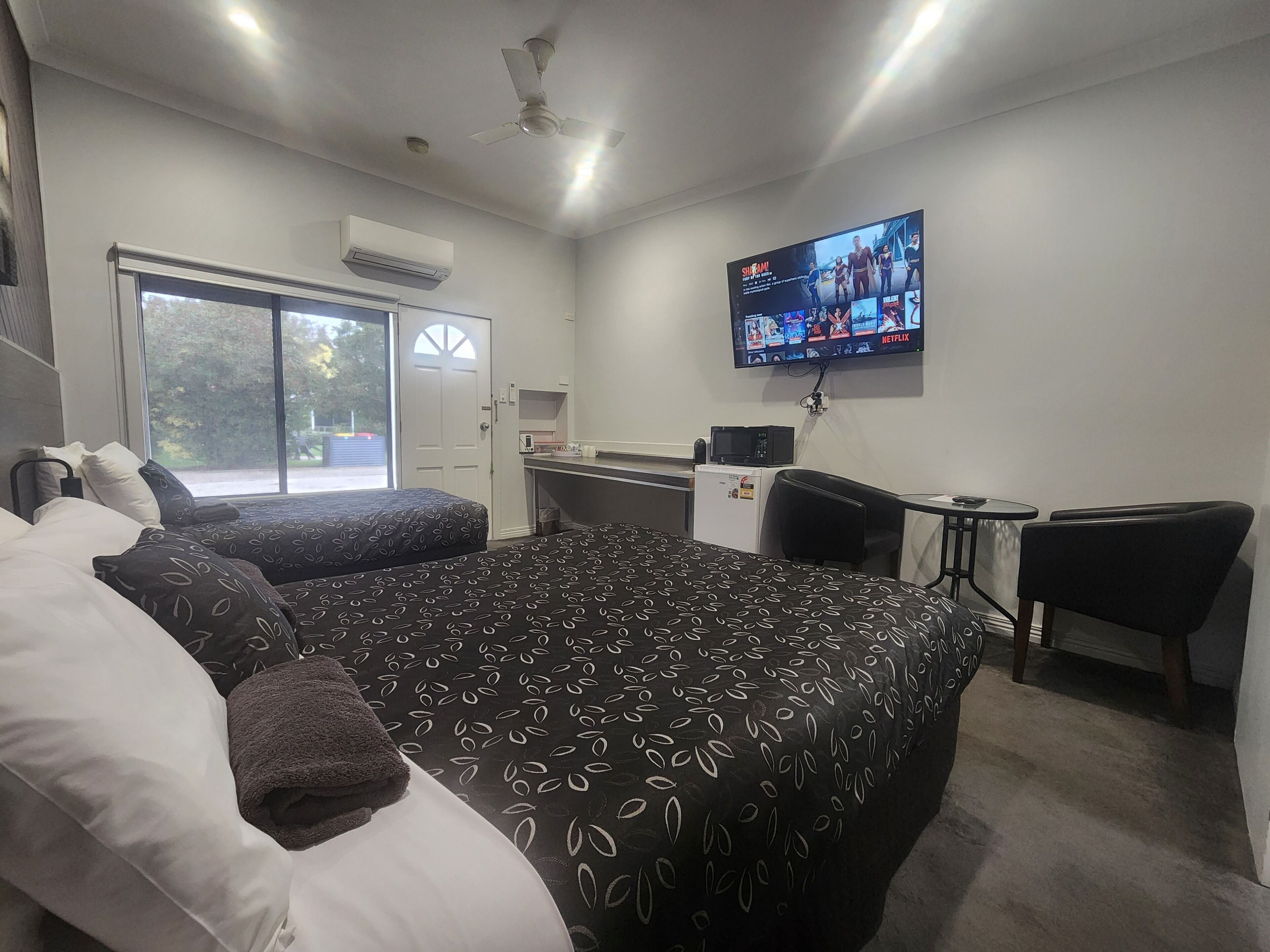 Habitación de hotel con una cama grande, un escritorio, una silla, un televisor montado en la pared y una ventana con vistas a árboles.
