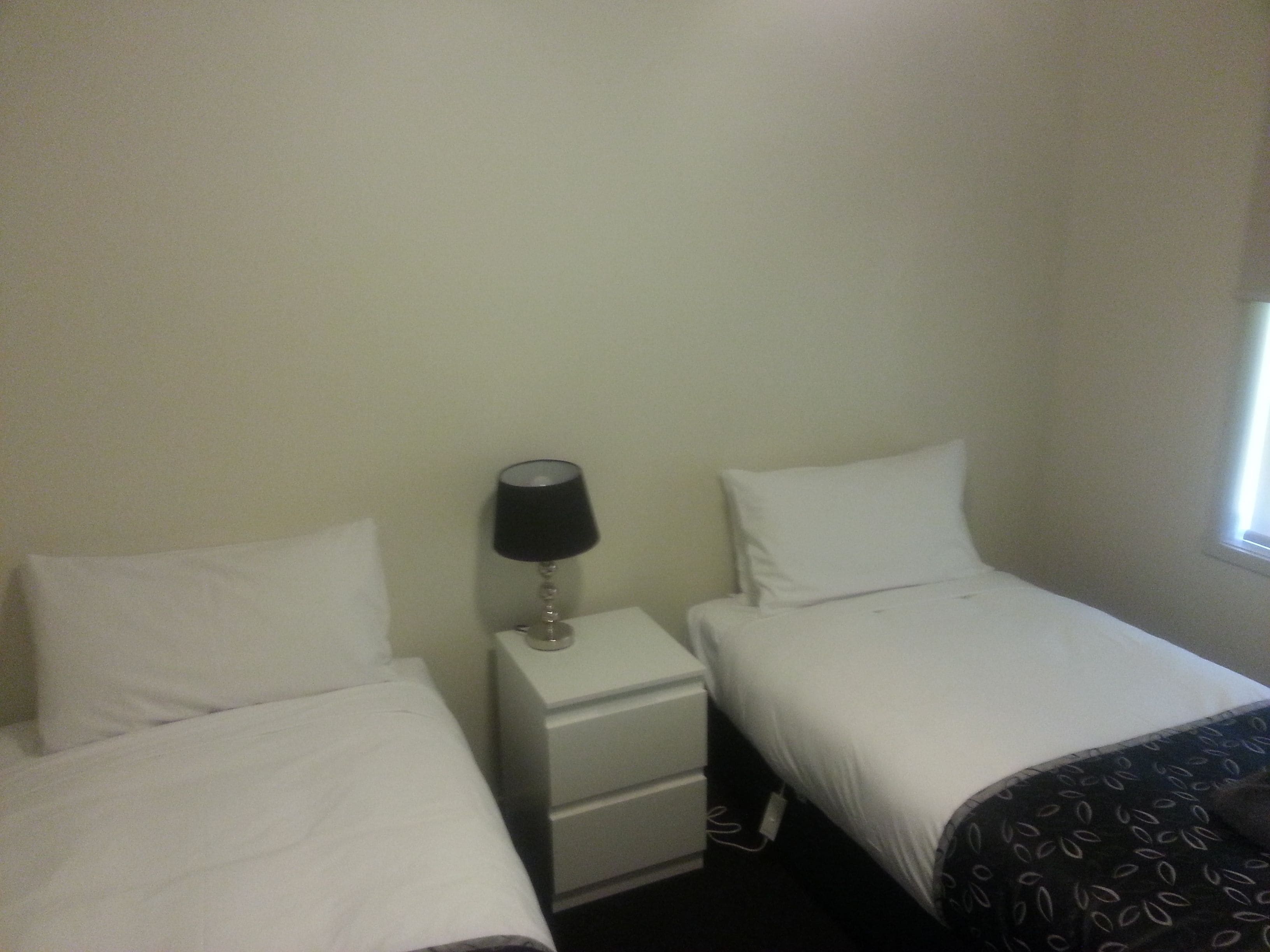 A small, simple room with two single beds, a nightstand with a lamp, and a window.