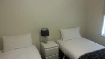 A small, simple room with two single beds, a nightstand with a lamp, and a window.