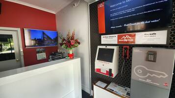 A hotel reception area with a red wall, a TV screen displaying a scenic view, a desk with a vase of flowers, and a signboard with hotel information and amenities.
