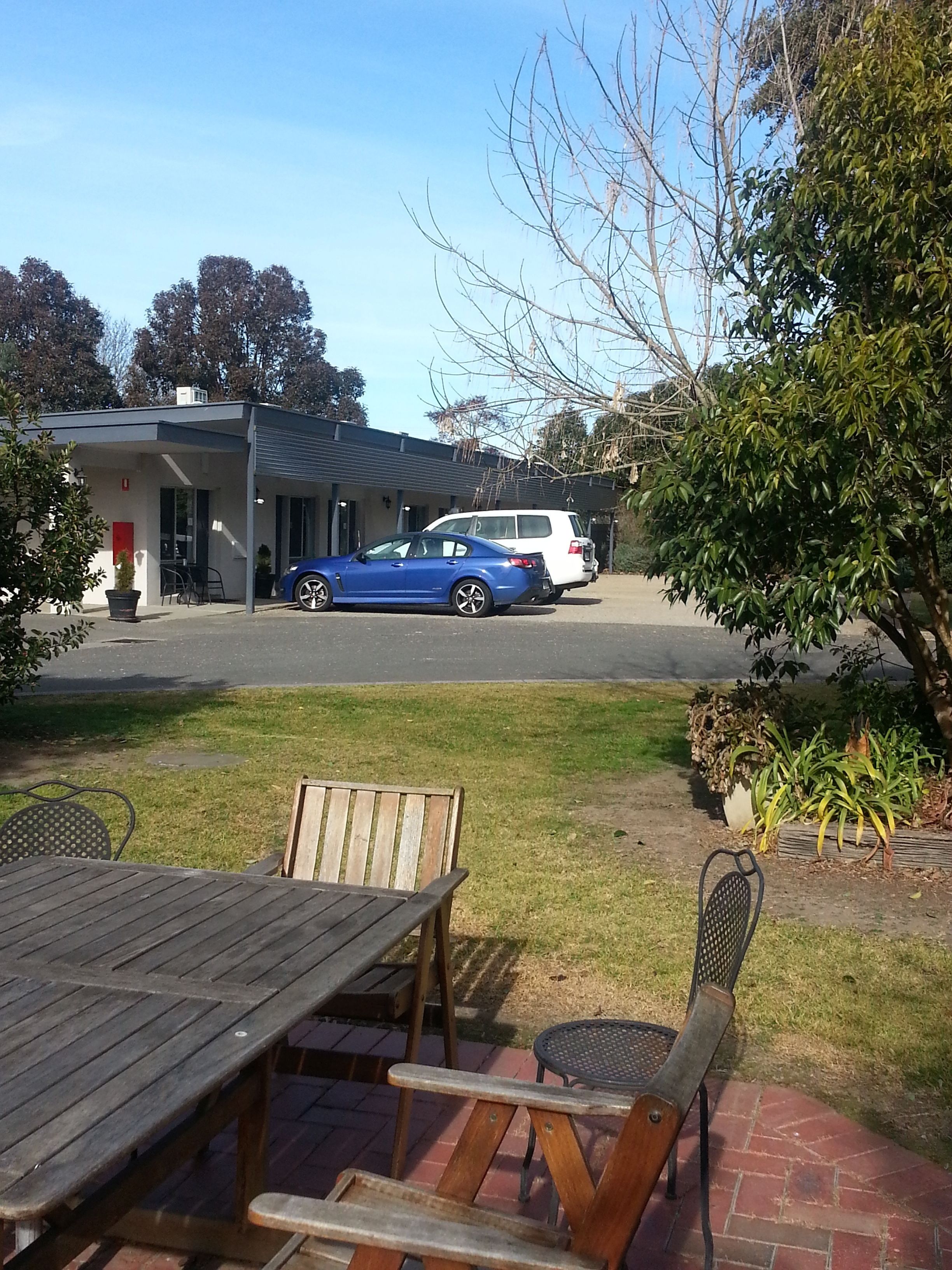 A parking area with a blue car and a white car parked in front of a building with a garage door. There are outdoor tables and chairs, and a grassy area with plants.