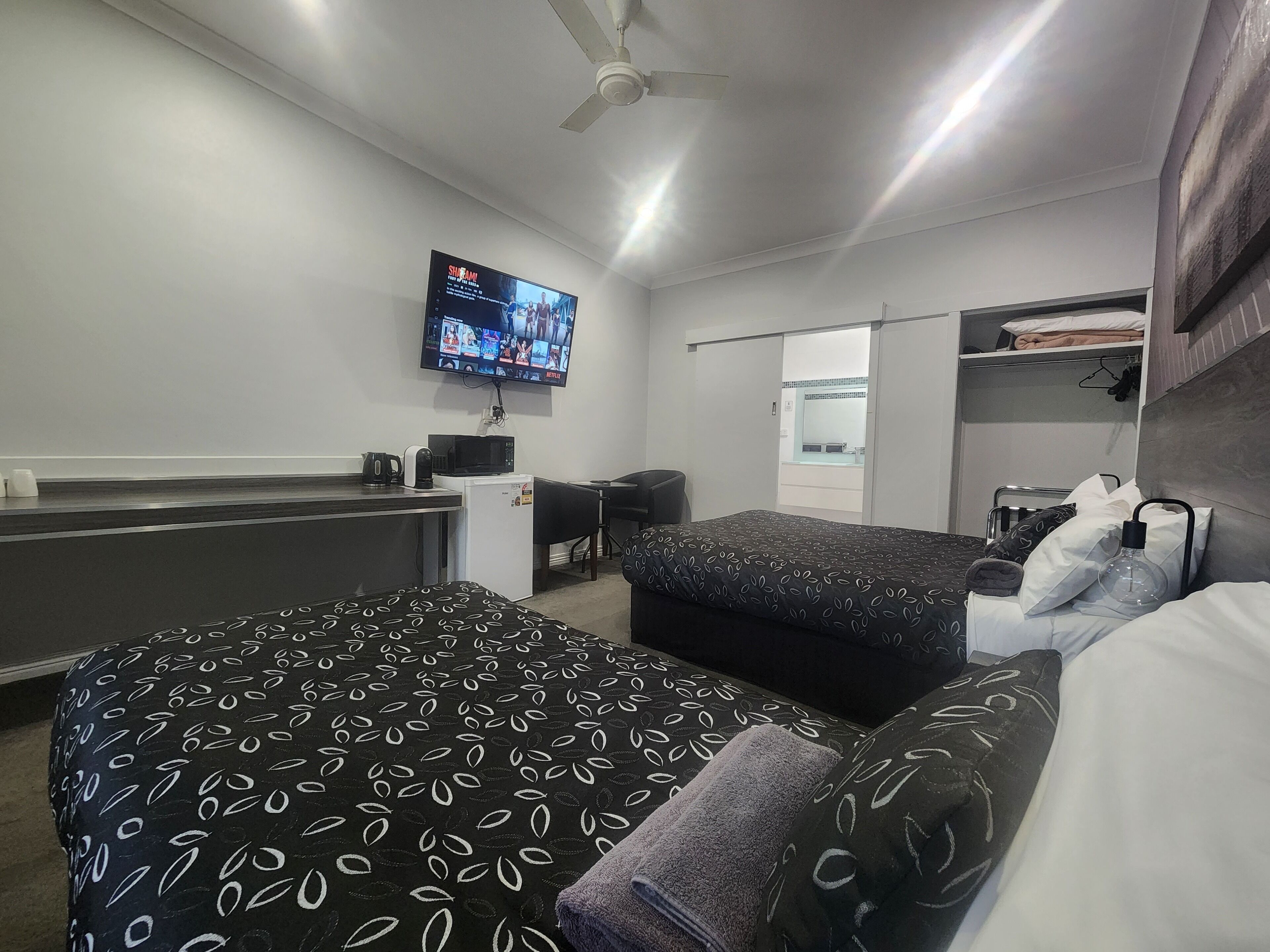 Habitación de hotel con dos camas, un escritorio, microondas, refrigerador, televisor mostrando un canal de deportes y ventilador de techo.