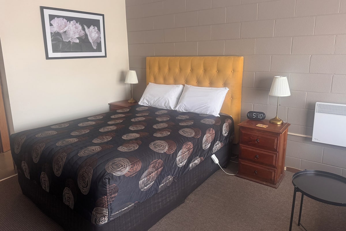 Una habitación de hotel con una cama, dos mesitas de noche, un reloj de pared, una lámpara y una imagen de flores.