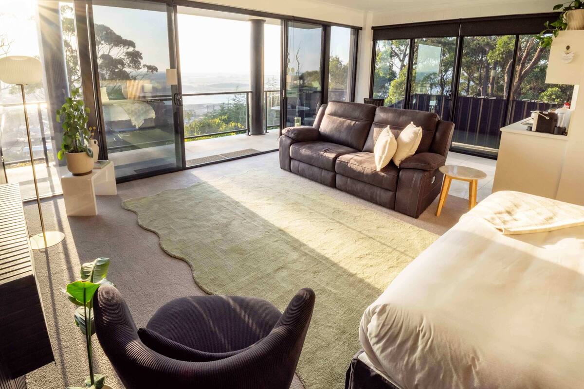 Quarto moderno com uma cama grande, um sofá de couro e vista do exterior através de janelas do chão ao teto.