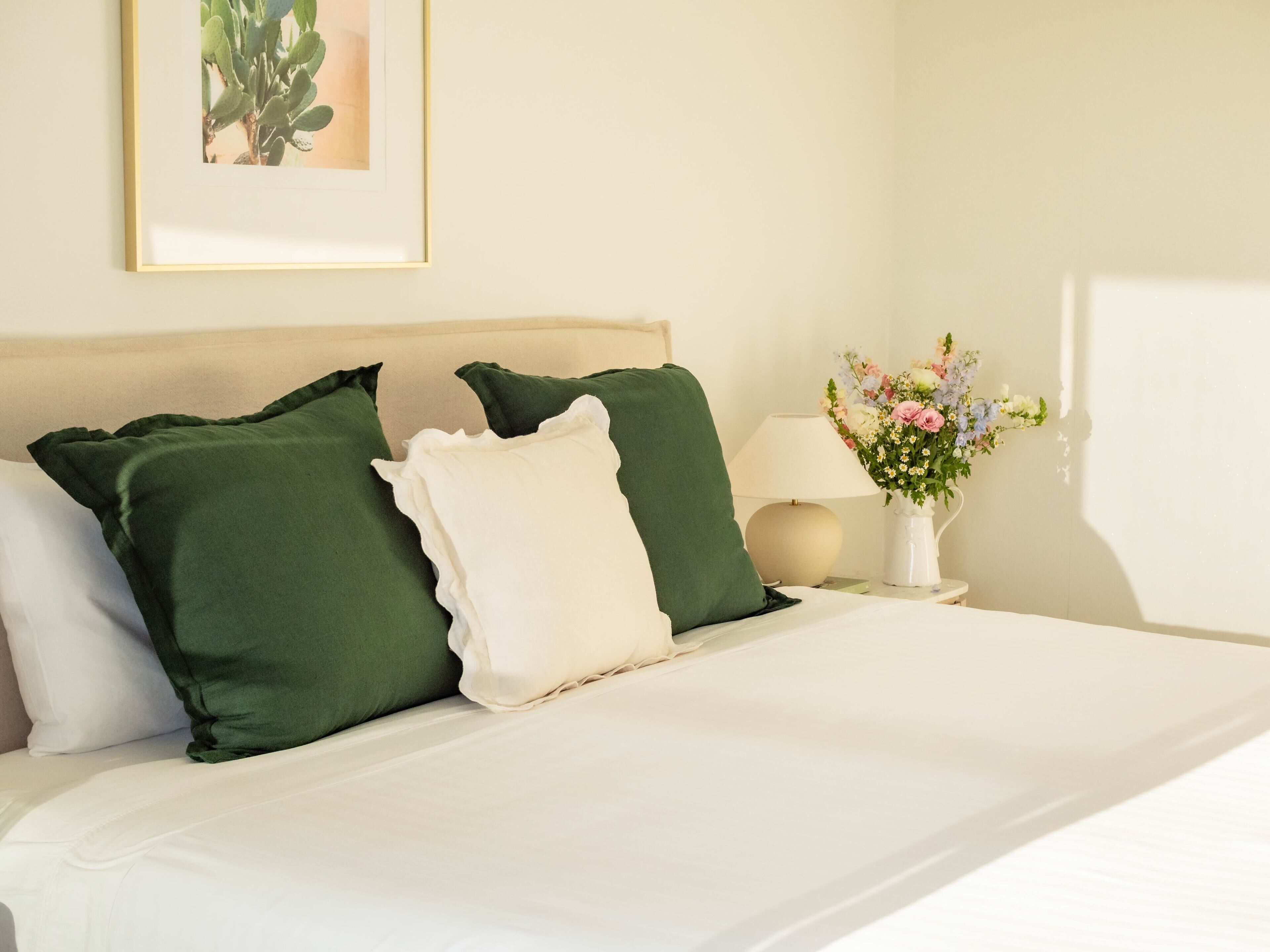 Una cama bien hecha con almohadas verdes y blancas, un cuadro en la pared, una mesita de noche con una lámpara y un florero con flores.