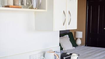 A hotel room with a bed, a bedside table with a coffee maker, a shelf with a kettle and glasses, and a wall socket.